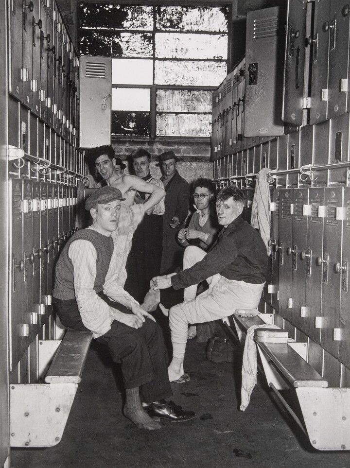 A black-and-white photograph of several men in various states of dress in the row of a locker room.