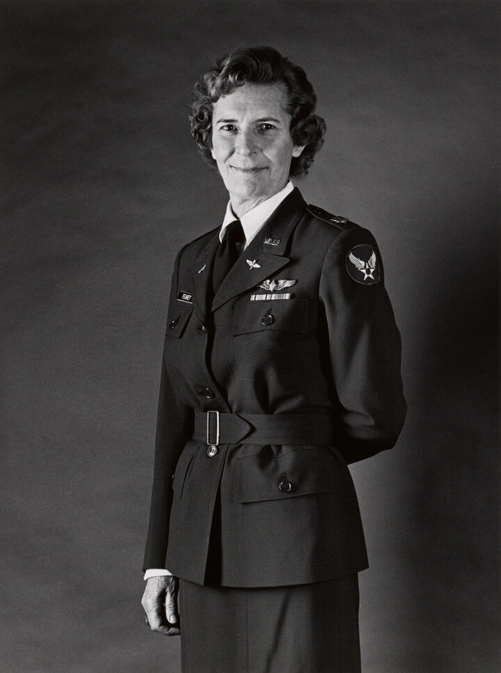 A black-and-white portrait photograph, three-quarter length, of a middle-aged White woman wearing a belted military jacket, one arm resting behind her back.