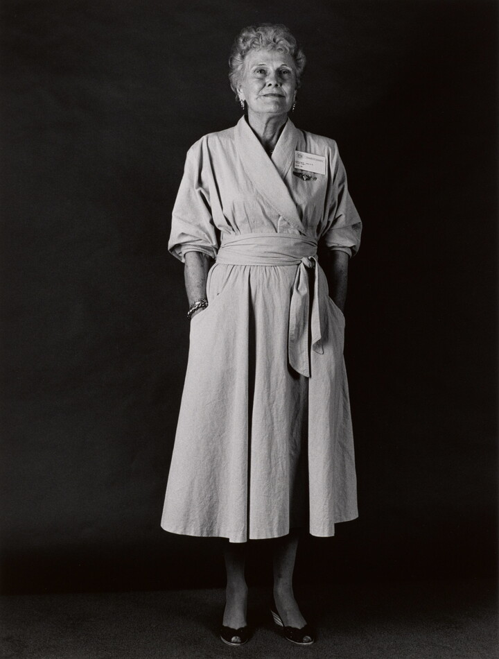 A black-and-white portrait photograph of an older White woman with short, light hair wearing a wrap dress with a name badge above her breast and her hands in her pockets.