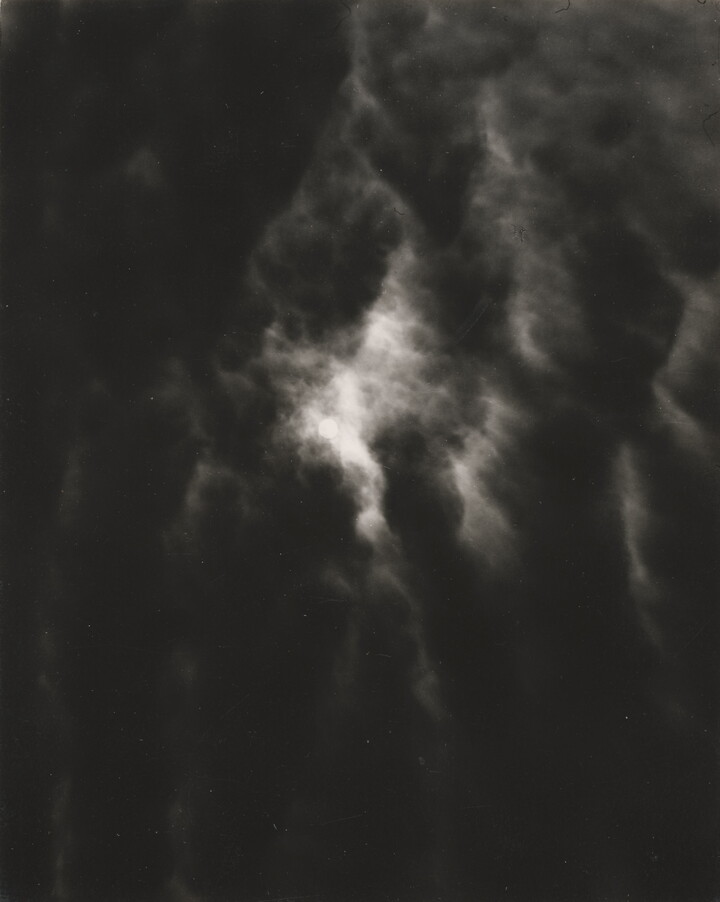 A black-and-white photograph of a dark, cloudy sky with light shining through the clouds.