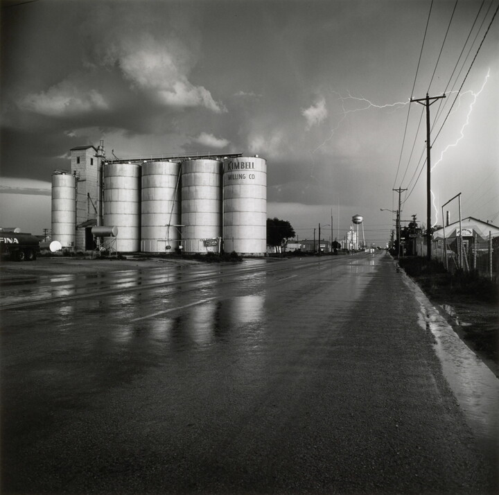 A black-and-white photograph of four grain elevators to the left of a wet road, a water tower in the far distance, and a flash of lighting behind power lines on the right.