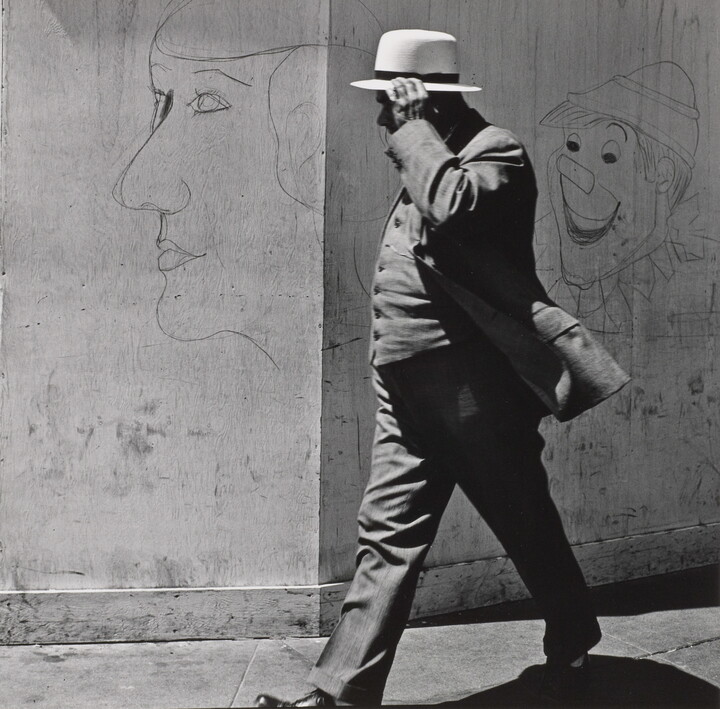 A black-and-white photograph of a man in a three-piece suit and hat walking past a wall covered with graffiti.