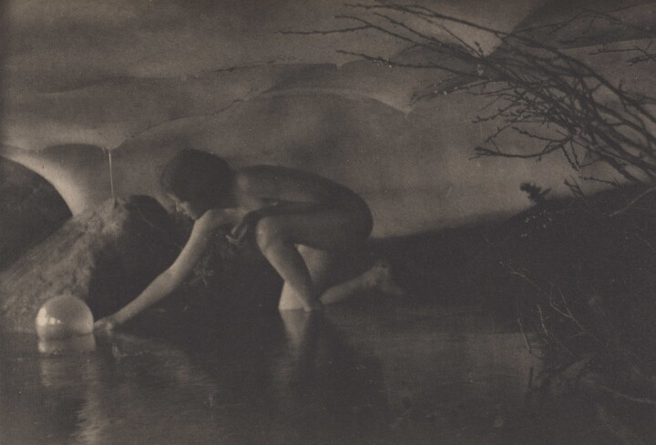A black-and-white photograph of a nude White women reaching down to pick up a sphere in a shallow body of water.