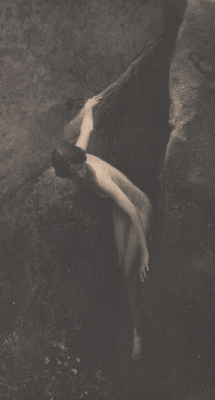A sepia-toned photograph of a nude White woman emerging from between two large boulders.