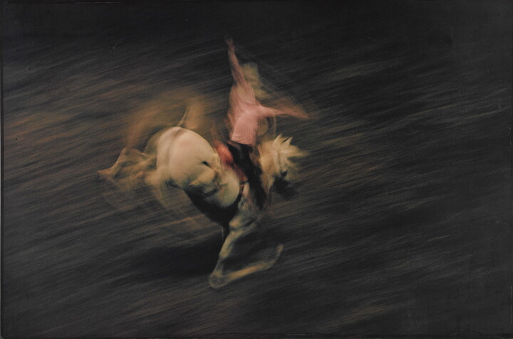 A color photograph of a cowboy on a bucking white horse, with the horse and cowboy blurred, indicating motion.