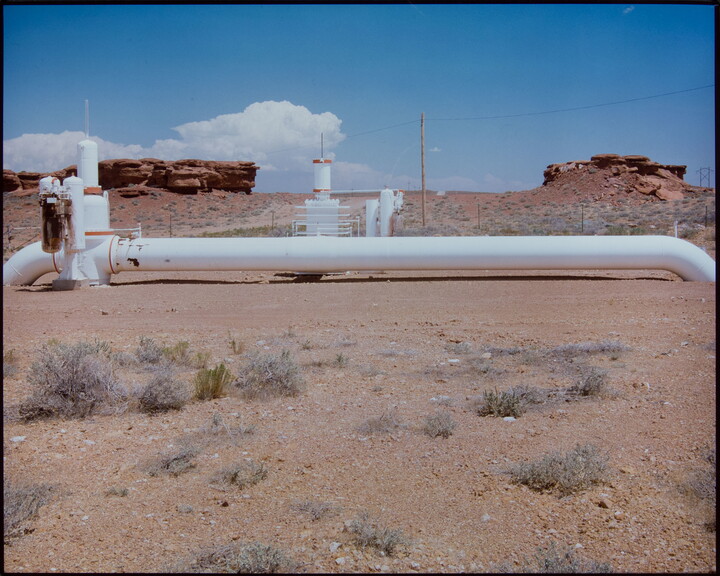 A color photograph of a white pipes and other machinery in a desert landscape with red rock formations in the background.