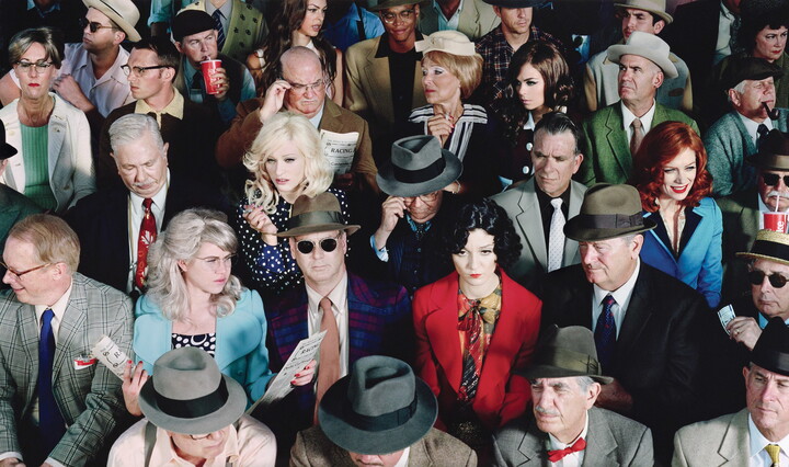 A color photograph of a crowd of people seated as if in a theater, most wearing brightly colored clothing and accessories.