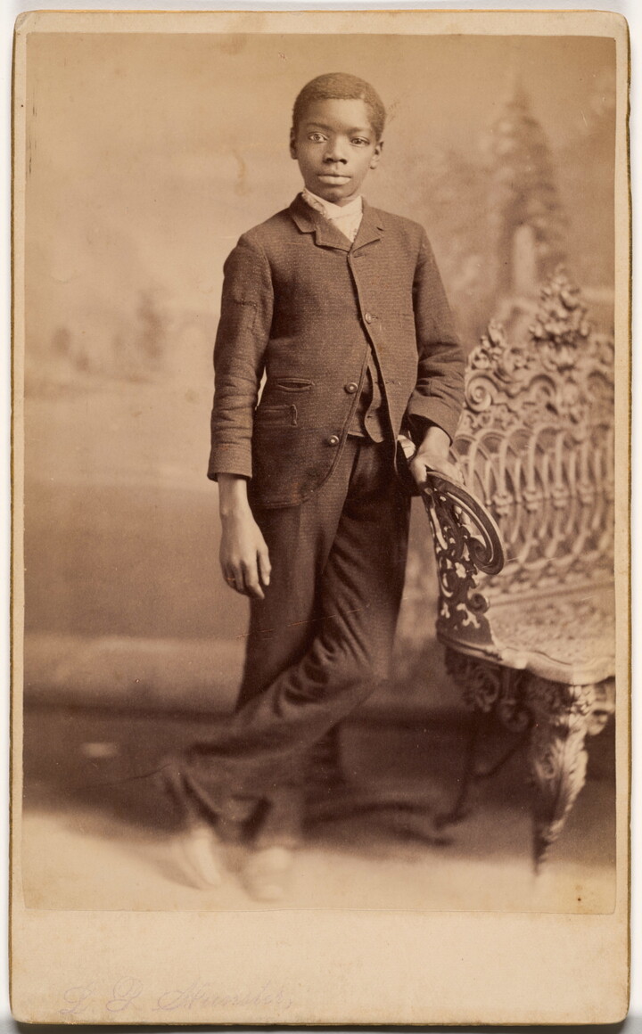 A sepia-toned studio portrait photograph of a Black boy wearing a suit, leaning against a wrought iron bench.