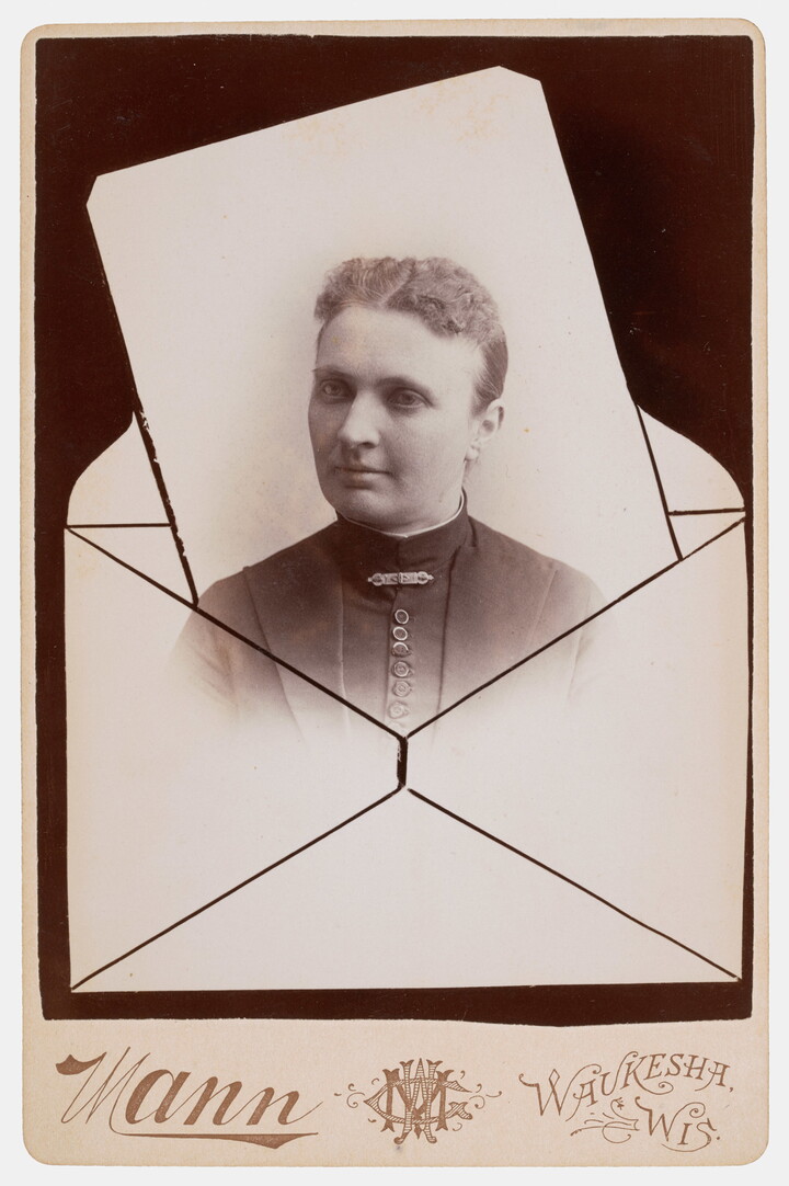 A sepia-toned studio photograph in which the head and shoulders of a White woman seems to be popping out of an envelope.