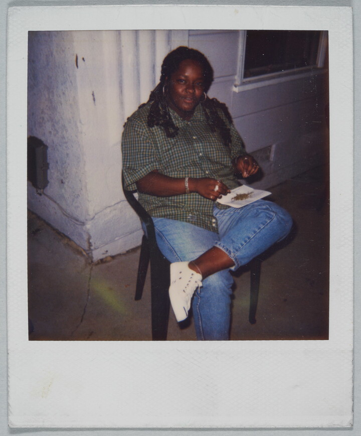 A color Polaroid photograph of a Black woman seated on a plastic chair balancing a piece of paper with bits of something on it on her crossed leg.