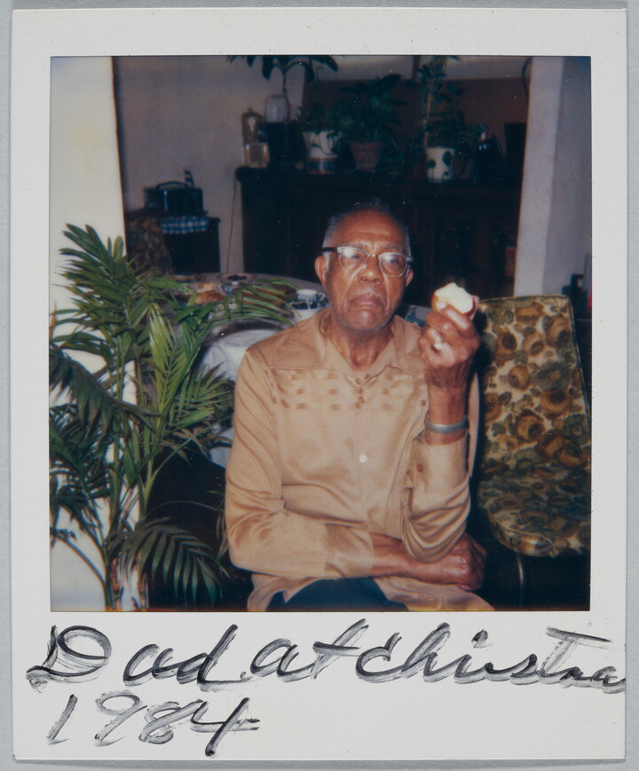 A color Polaroid photograph of a seated, middle-aged Black man in glasses and a light brown shirt holding an apple in one hand; handwritten text at bottom reads: "Dad at Christma[s] 1984."