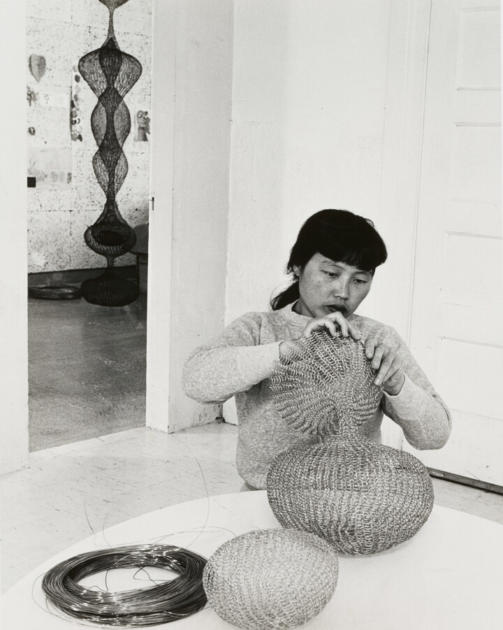 A black-and-white photograph of a woman with Asian features seated at a table and working an a wire sculpture.