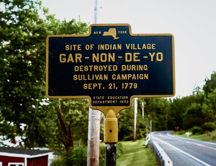 A color photograph of a New York historical marker that reads, "Site of Indian Village Gar-Non-De-Yo Destroyed during Sullivan Campaign Sept. 21, 1779," next to a road.