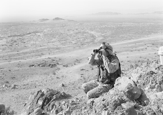 A black-and-white photograph of a man dressed in military fatigues kneeling in a rocky, desert landscape looking through binoculars.
