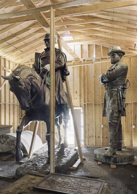 A color photograph of two bronze sculptures, one of a man on horseback and the other of a standing man in military uniform, in a barn-like structure.