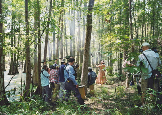 A color photograph of a film crew in a thickly wooded swamp filming a White woman in 19th-century clothing aiming a rifle.