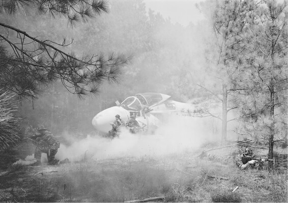 A black-and-white photograph of soldiers in fatigues in a wooded setting surrounding an airplane on the ground.