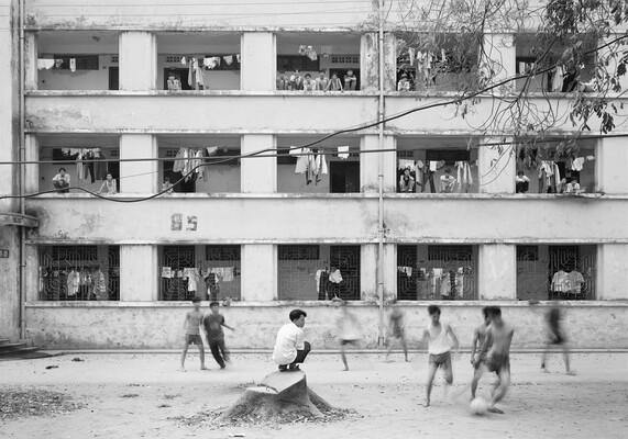 A black-and-white photograph of a group of young Asian men playing soccer in a dirt courtyard of a multi-story apartment building.