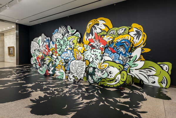 An installation photo of large wood cut-outs of flowers and leaves, some brightly-colored, against a black wall with shadows projected on the floor in a Carter gallery. 