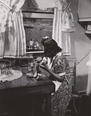 A black-and-white photograph of a Black woman seated at a kitchen table peeling potatoes as she watches children children in the yard through a window.