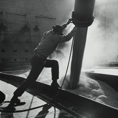 A black-and-white photograph of a man holding a pipe that is shooting grain onto a platform.