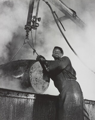 A black-and-white photograph of a Black man wearing a long apron holding a large barrel that is suspended by a piece of machinery.