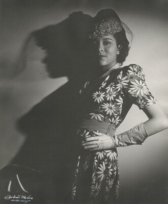 A black-and-white photograph of a Black woman wearing a pillbox hat with a net veil and a dress with a floral pattern, one gloved hand on her hip, leaning against a wall.