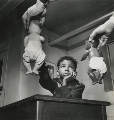 A black-and-white photograph of a Black child seated at a desk as someone out of the frame holds out two baby dolls - one White and one Black - and the child reaches toward the White one.