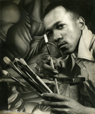 A black-and-white photograph of a close-up of a Black man holding several paintbrushes in front a mural.
