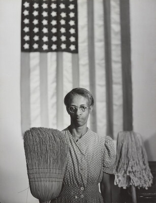 A black-and-white photograph of a middle-aged Black woman wearing a housedress, an American flag hanging behind her, and an upturned broom and mop in front of her.