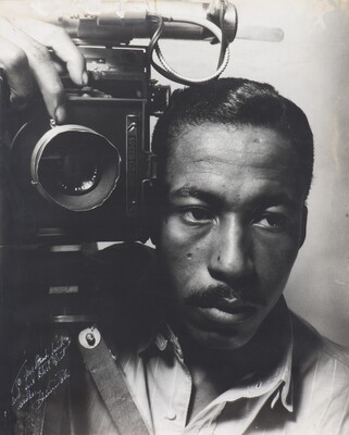 A black-and-white photograph of a Black man with a mustache holding a large camera on his right shoulder.