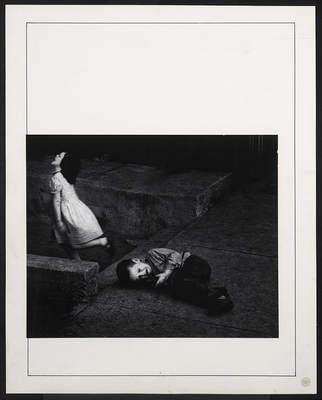 A black-and-white photograph of two White children: a girl in a white dress running from a boy who lies on a concrete sidewalk crying.
