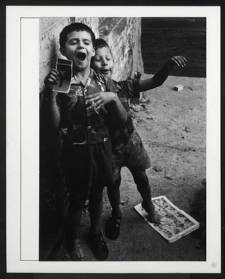 A black-and-white photograph of two young White boys standing next to each other against a brick wall; the taller boy holds photos in his hand and the other gestures with his arms.