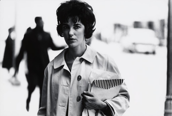 A black-and-white photograph of a White woman with short dark hair, wearing a raincoat, and holding large envelopes in her arm.
