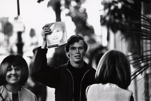 A black-and-white photograph of three people on a sidewalk: a smiling White woman with dark, bobbed hair and bangs; a smiling White man holding up a book with a drawing of a woman's face on the book cover; the back of another person. 