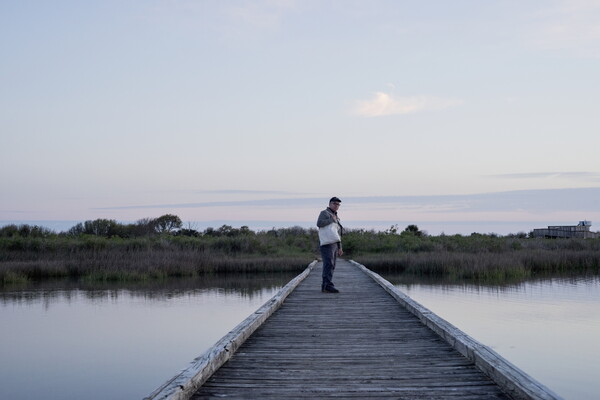 A color photograph of a White man standing on a wood boardwalk that goes over water.