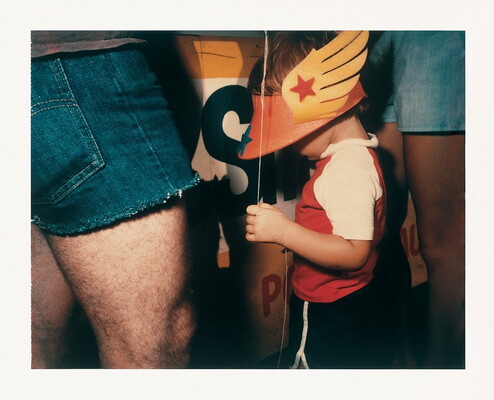 A close-cropped color photograph of the hips and thigh of a White adult next to a White child wearing an orange sun visor with yellow wings and a balloon string around their wrist.