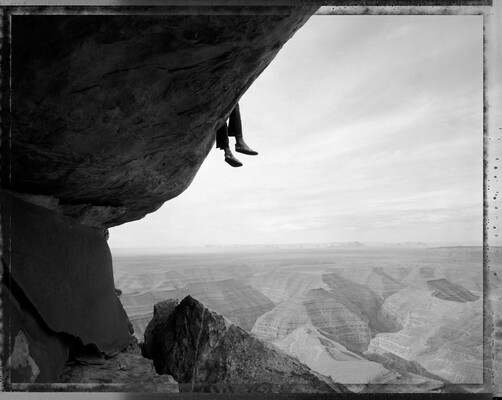 A black-and-white photograph taken from under a cliff ledge showing feet dangling over with canyon rock formations in the distance.