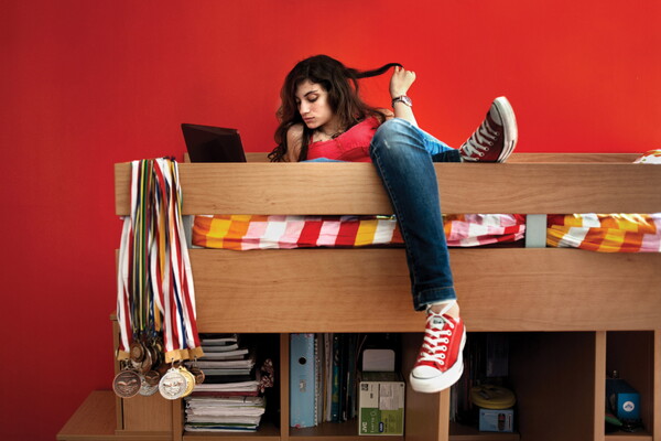 A color photograph of a light-skinned teenage girl lounging on a top bunk, twirling her long hair around a finger, sports medals slung around a bedpost.