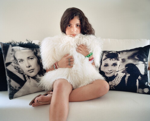 A color photograph of a light-skinned pre-teen girl seated on a white sofa holding a fuzzy white pillow in front of her torso.