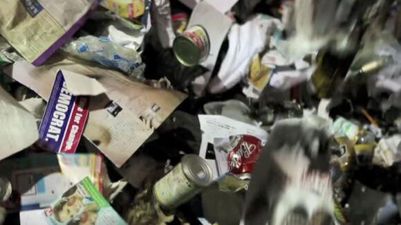 A video still of garbage, including paper, cans, plastic bags.