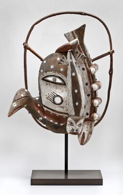 A carved wood mask that features animal faces or heads in white and red, and what looks like human fingers curling around the edge.