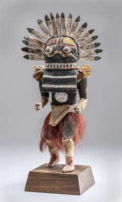 A wood sculpture of a human-like figure posed as if it were dancing; a fan of feathers across its head and a red grass-like skirt around its waist.