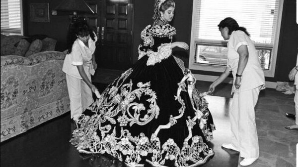 A black-and-white photograph of a young White woman wearing a formal gown with a full skirt as two women in white help arrange her skirt.