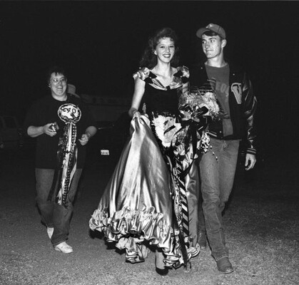 A black-and-white photograph of a smiling teenage White girl wearing a formal gown walking arm-in-arm with a White teenage boy wearing a ball cap and a jacket.