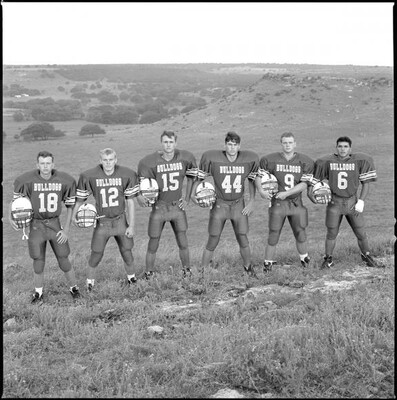 A black-and-white photograph of six White high school football players, in their uniforms, standing on a grassy hillside.
