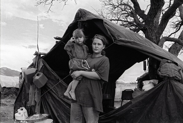 A black-and-white photograph of a young White woman holding a young child on her hip standing in front of a canvas tent under a bare tree.