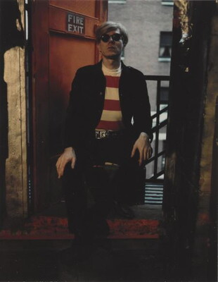 A color photograph of a White man with short blond hair, dark sunglasses, a red and white striped shirt under a black jacket, and black pants sitting in front of a red door with a sign that reads, "Fire Exit."