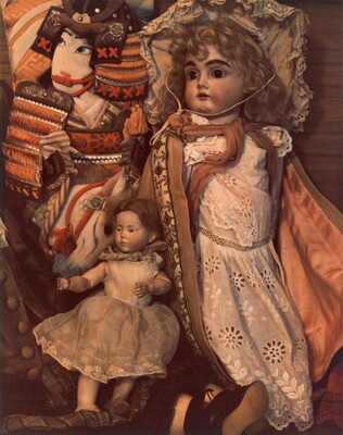 A color photograph of a pile of dolls: a quilted doll with Asian features, a china doll with blonde curly hair, and a smaller doll in a white dress.