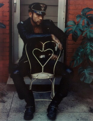 A color photograph of a White man, dressed all in black and wearing a black cap with a skull and crossbones on the front panel, sitting backward in a Victorian-style wire chair.